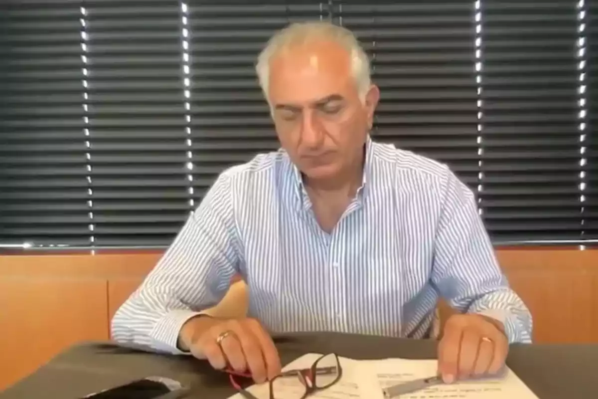 Hombre de cabello canoso y camisa de rayas sentado en una mesa revisando documentos con unas gafas y un bolígrafo en la mano frente a una persiana negra Hombre de cabello canoso y camisa de rayas sentado en una mesa revisando documentos con unas gafas y un bolígrafo en la mano frente a una persiana negra