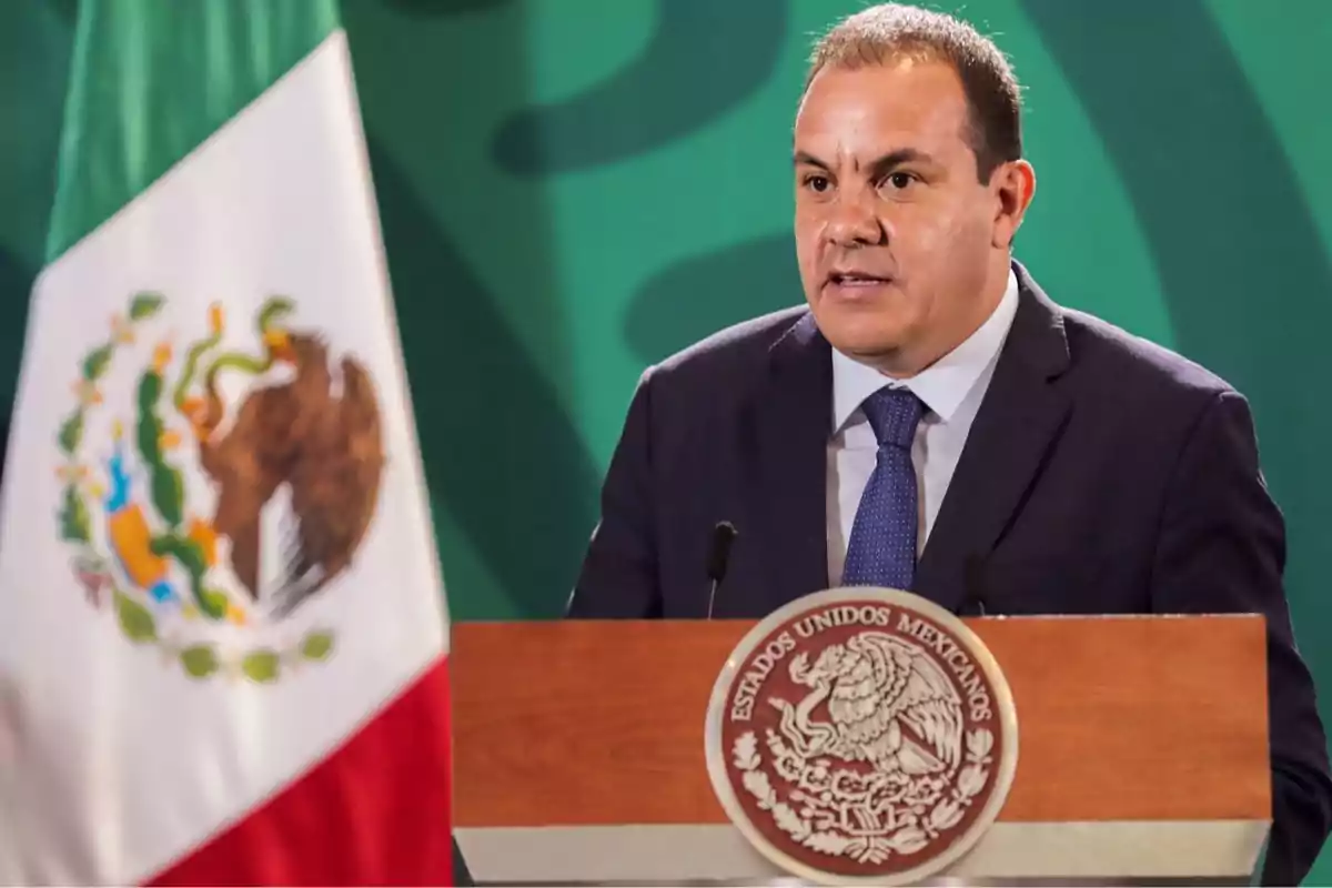 Un hombre en traje habla en un podio con el escudo de México, junto a una bandera mexicana.