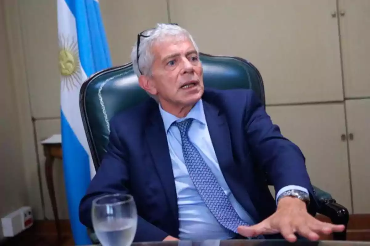 Un hombre de cabello canoso y traje azul está sentado en una silla de oficina con una bandera argentina de fondo.