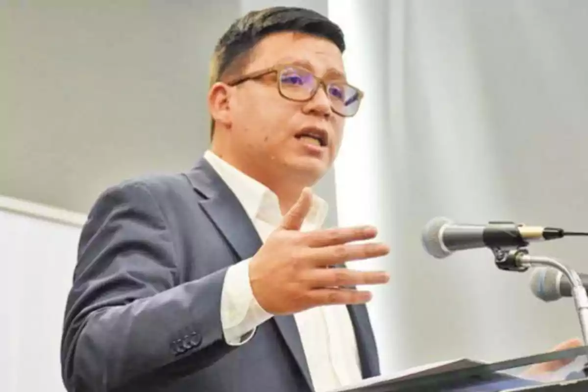 Man with glasses and a blue blazer speaking in front of a microphone at a podium Man with glasses and a blue blazer speaking in front of a microphone at a podium