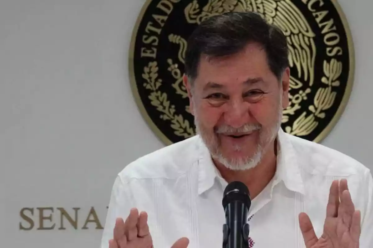 Gray-bearded man in a white jersey smiling as he speaks into a microphone with the national emblem of Mexico in the background Gray-bearded man in a white jersey smiling as he speaks into a microphone with the national emblem of Mexico in the background