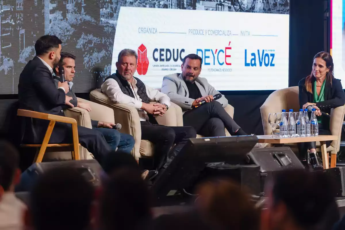 Cinco personas participan en un panel de discusión en un escenario, sentados en sillones y conversando frente a una audiencia, con botellas de agua sobre una mesa y un fondo con logotipos de empresas.