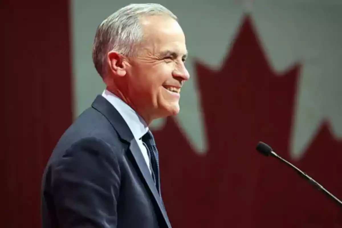 Hombre de cabello canoso con traje oscuro sonriendo frente a un micrófono con una bandera de Canadá de fondo Hombre de cabello canoso con traje oscuro sonriendo frente a un micrófono con una bandera de Canadá de fondo