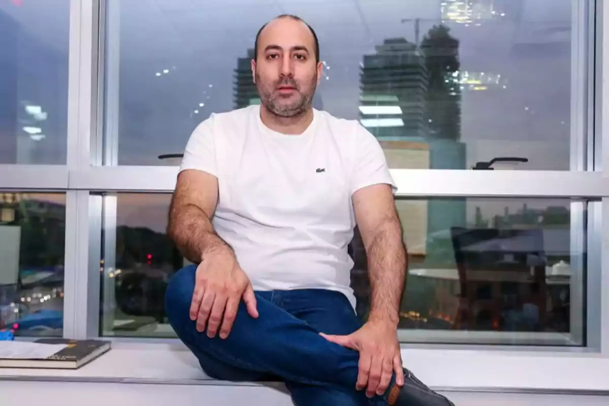 Hombre sentado en el alféizar de una ventana con edificios al fondo.