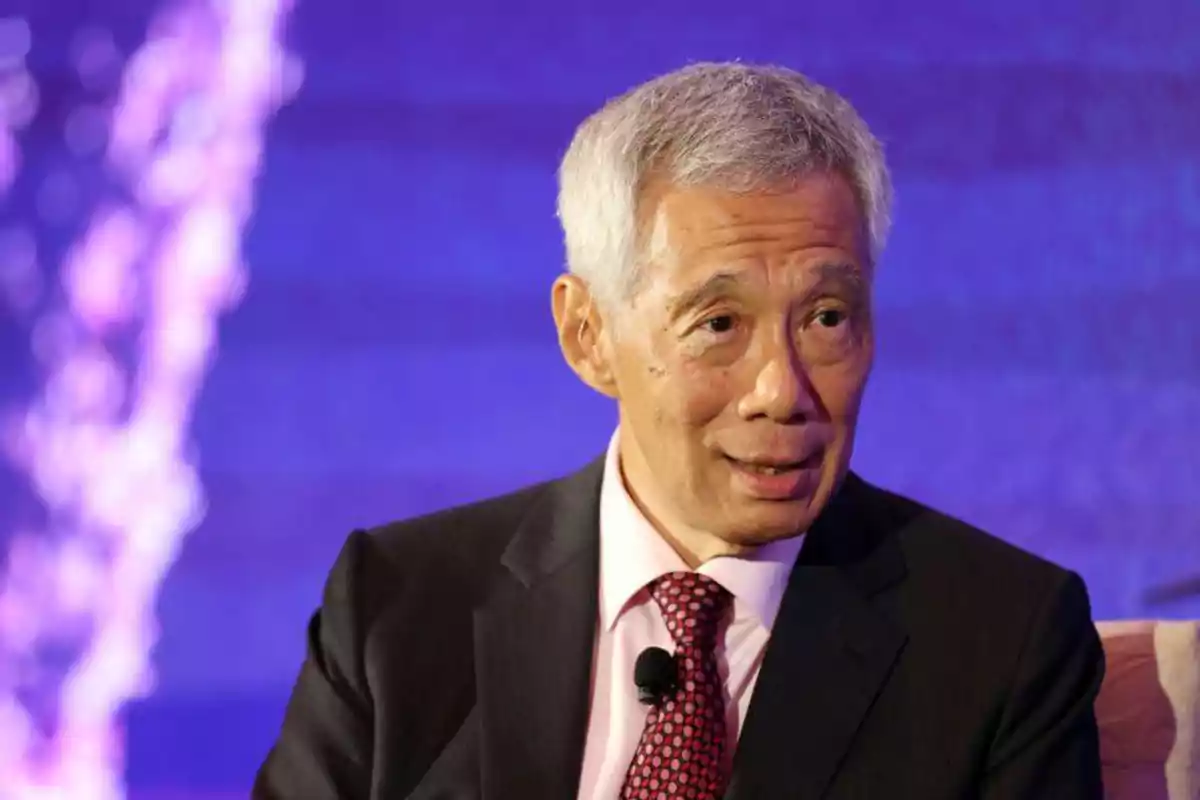 A gray-haired man in a dark suit with a red tie speaks at an event with a blue background. A gray-haired man in a dark suit with a red tie speaks at an event with a blue background.