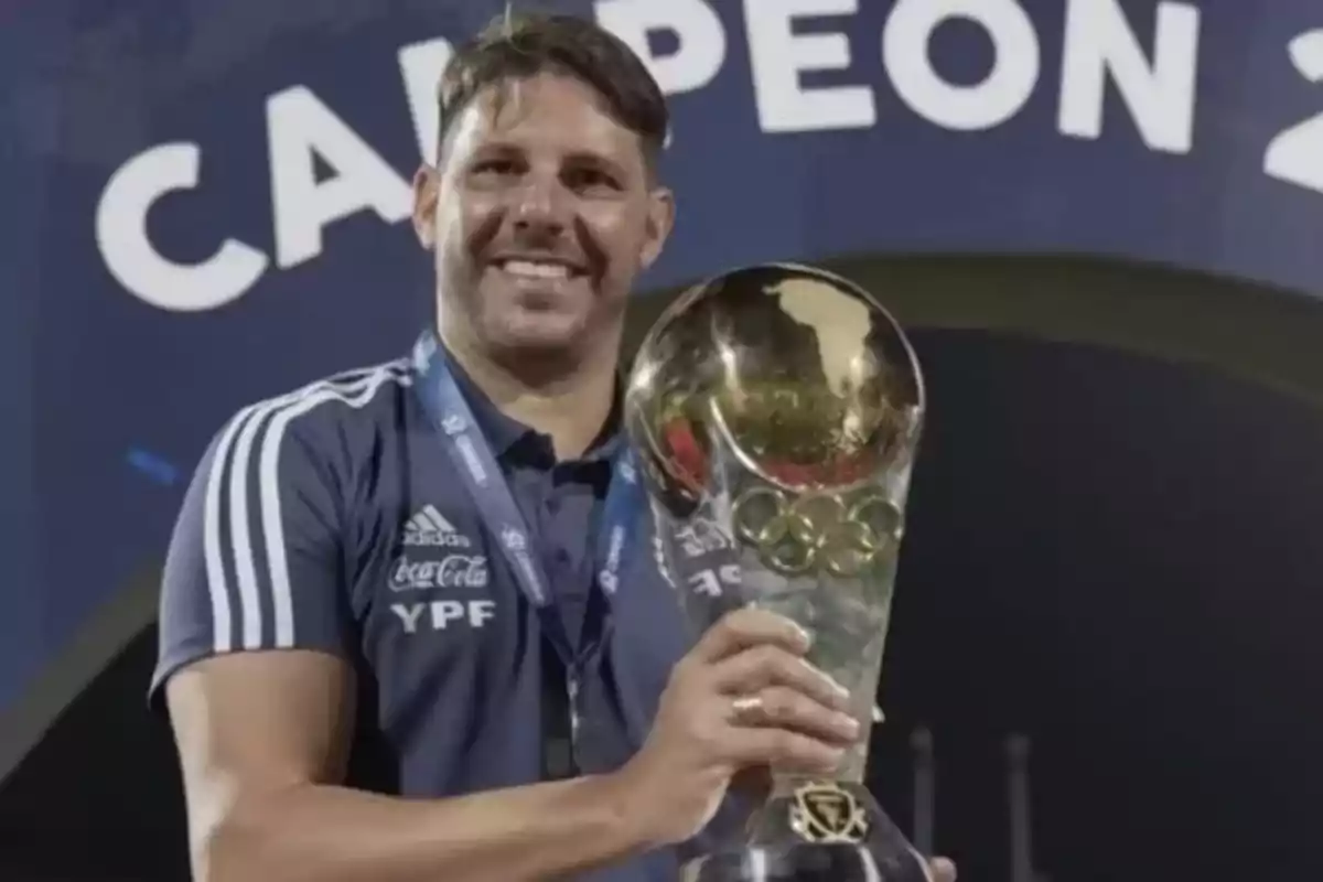 Smiling man in a blue sports jersey holds a golden trophy in front of a champion banner Smiling man in a blue sports jersey holds a golden trophy in front of a champion banner
