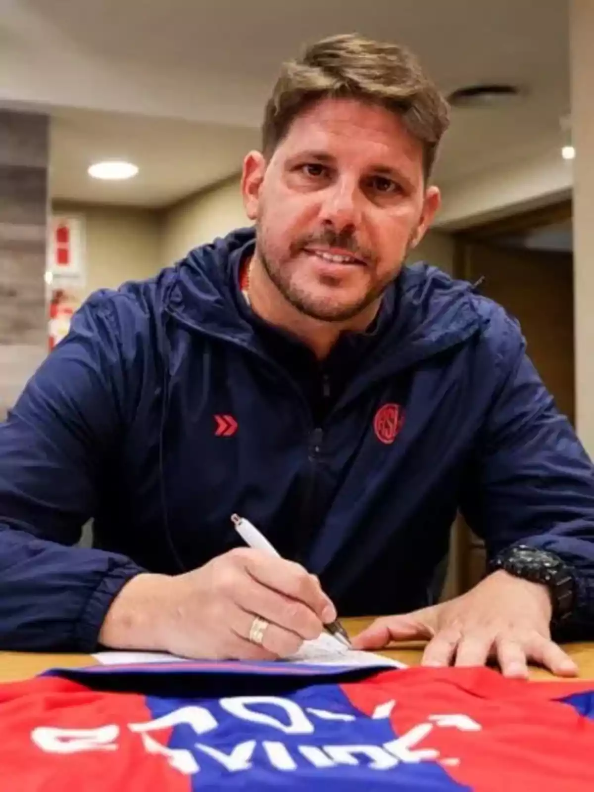 Hombre con chaqueta azul firmando un documento sobre una mesa donde hay una camiseta deportiva roja y azul Hombre con chaqueta azul firmando un documento sobre una mesa donde hay una camiseta deportiva roja y azul