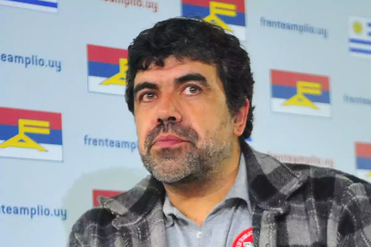 Dark-haired, bearded man with a serious expression in front of a background with logos and flags of Uruguay’s Broad Front.