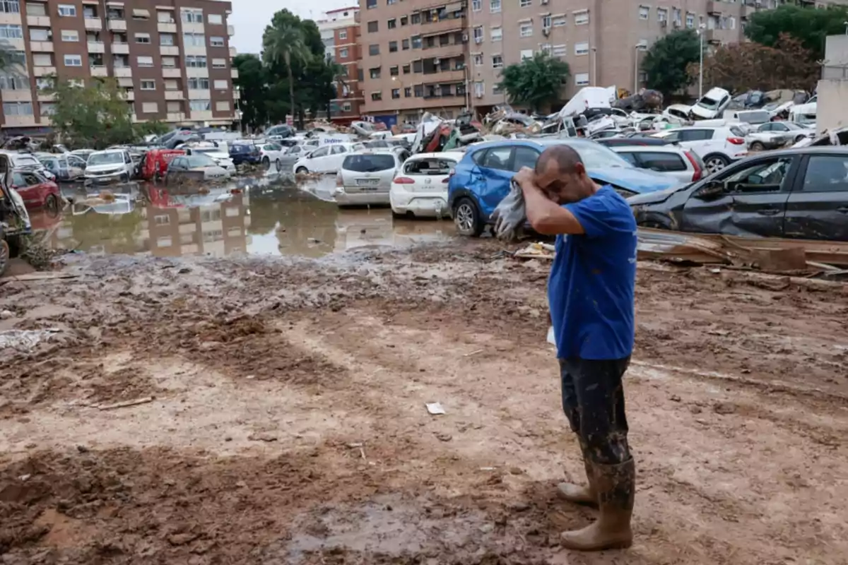 Un hombre se seca el sudor en una zona inundada y llena de barro con coches dañados y edificios al fondo.