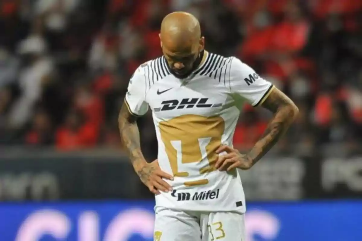 Jugador de fútbol con uniforme blanco y dorado mirando hacia abajo en un campo de juego.