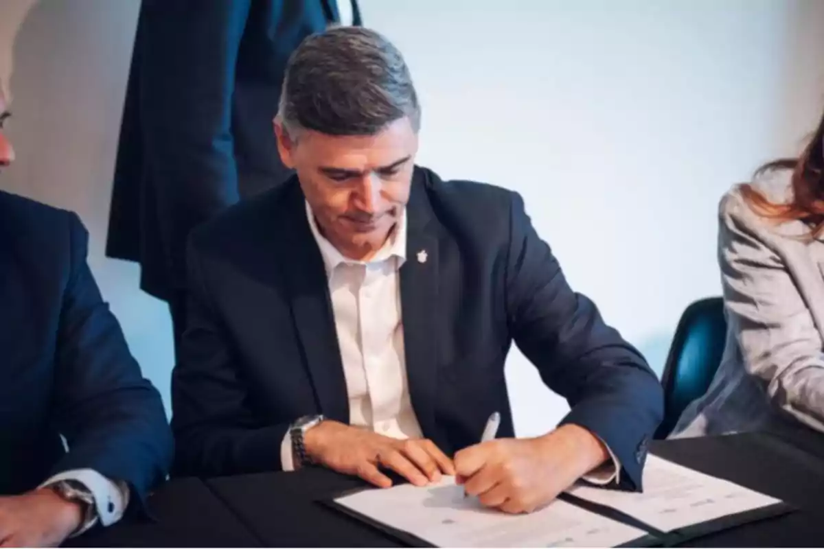 Hombre firmando un documento en una mesa junto a otras personas.