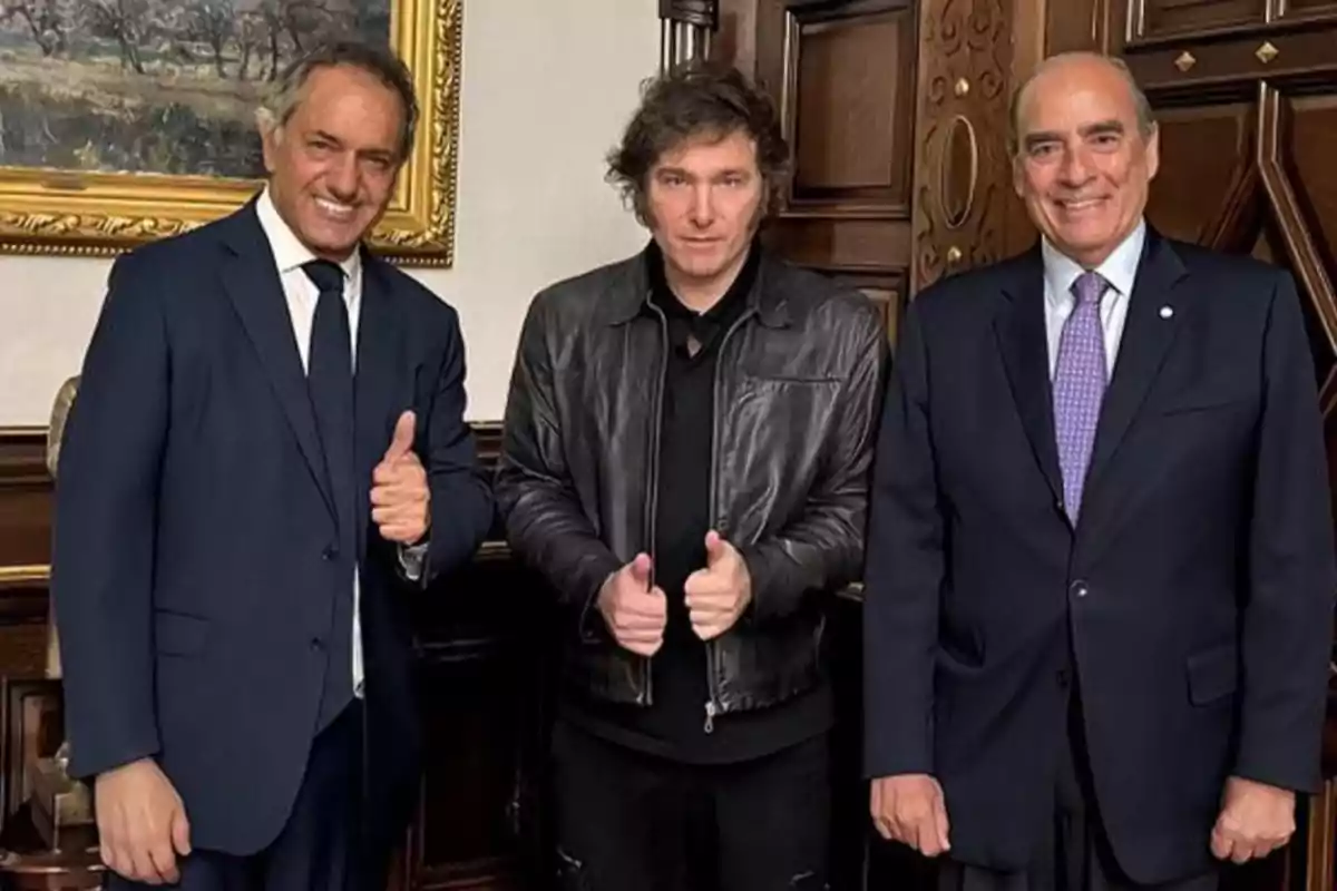 Tres hombres posan juntos en un ambiente formal, dos de ellos llevan traje y corbata mientras el del centro viste una chaqueta de cuero negra y todos sonríen a la cámara Tres hombres posan juntos en un ambiente formal, dos de ellos llevan traje y corbata mientras el del centro viste una chaqueta de cuero negra y todos sonríen a la cámara
