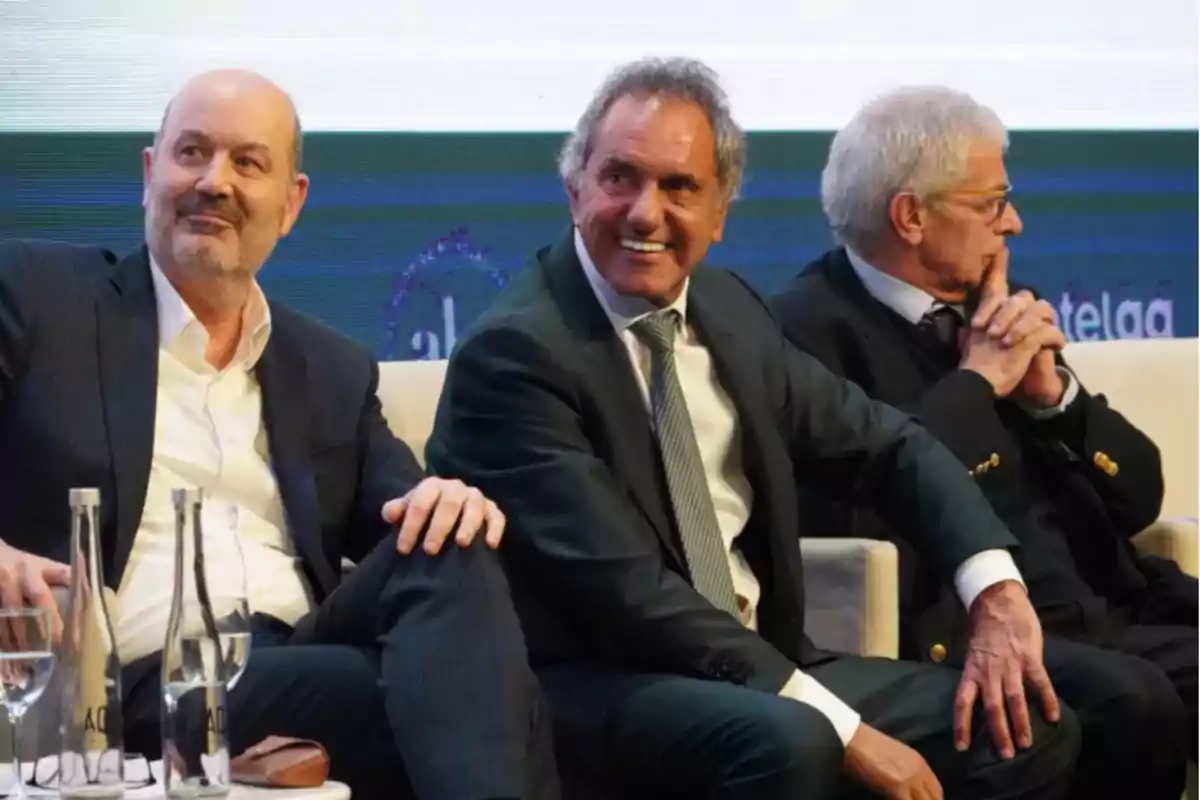 Tres hombres sentados en un panel de discusión, todos con trajes formales y sonriendo.