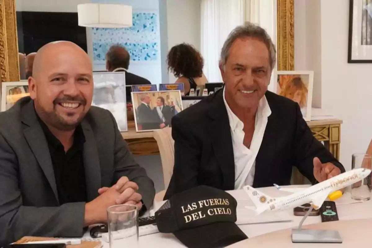 Dos hombres sonríen sentados en una mesa con una gorra negra que dice Las Fuerzas del Cielo y un modelo de avión frente a ellos, al fondo hay fotos enmarcadas y un espejo grande.
