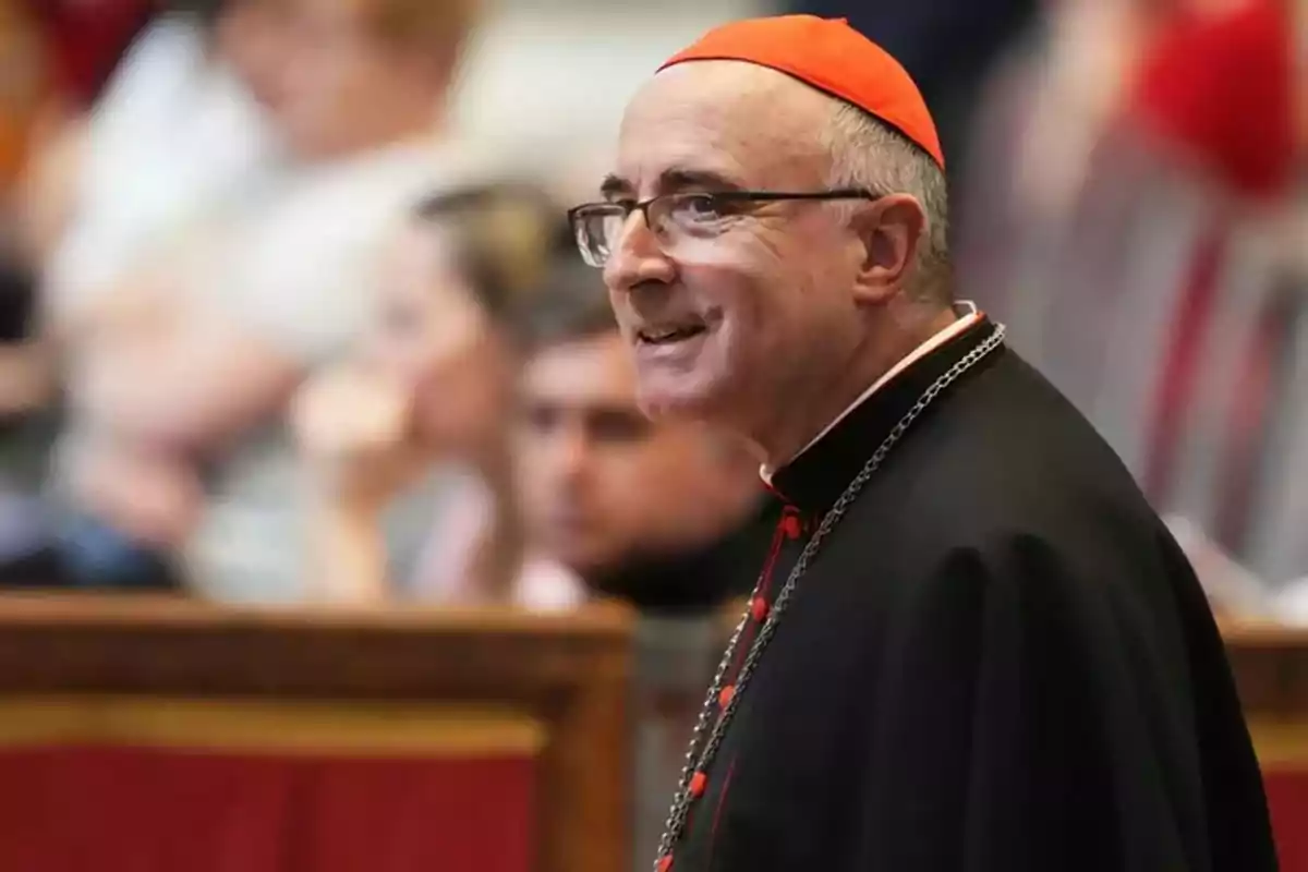 A cardinal with a red biretta and black ecclesiastical attire smiles while standing in a place with people in the background. A cardinal with a red biretta and black ecclesiastical attire smiles while standing in a place with people in the background.