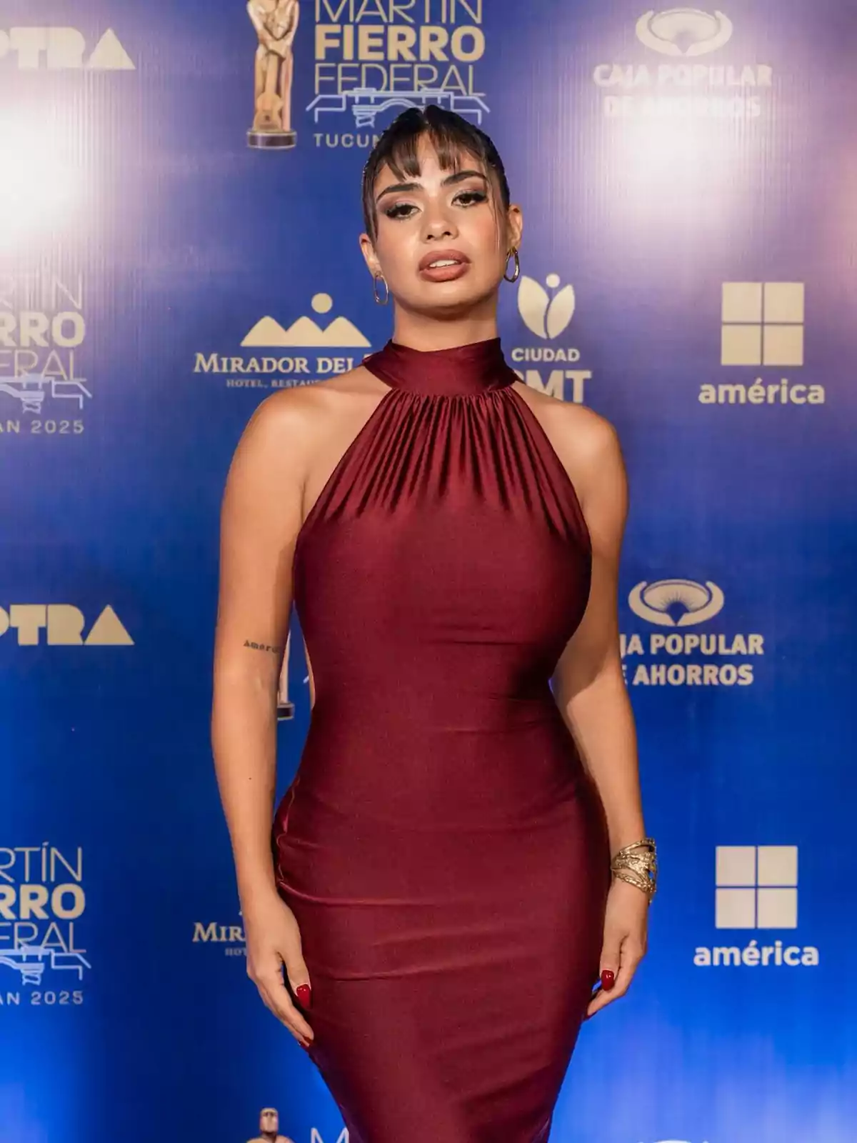 A person in a burgundy dress poses in front of a blue background with event logos. A person in a burgundy dress poses in front of a blue background with event logos.