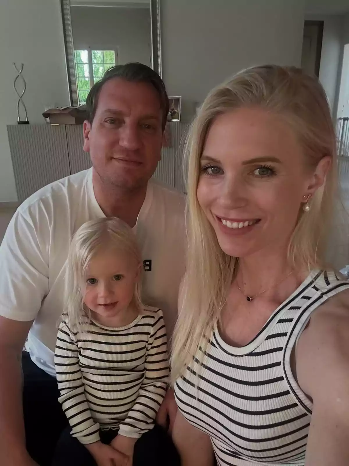 Una familia sonriente posa para una foto en el interior de una casa, la madre y la hija llevan camisetas de rayas blancas y negras y el padre viste una camiseta blanca. Una familia sonriente posa para una foto en el interior de una casa, la madre y la hija llevan camisetas de rayas blancas y negras y el padre viste una camiseta blanca.