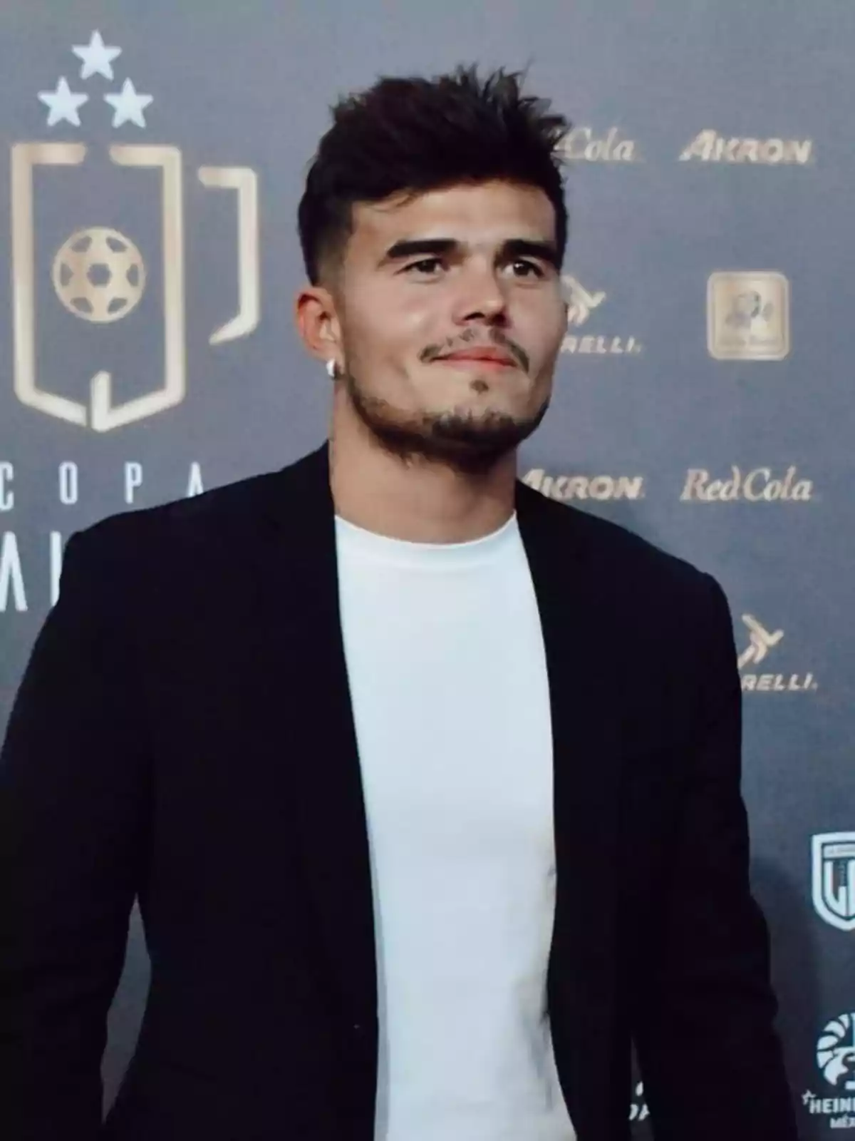 A man with a beard and short hair, wearing a white t-jersey and a black jacket, posing in front of a background with logos and a stylized soccer trophy. A man with a beard and short hair, wearing a white t-jersey and a black jacket, posing in front of a background with logos and a stylized soccer trophy.