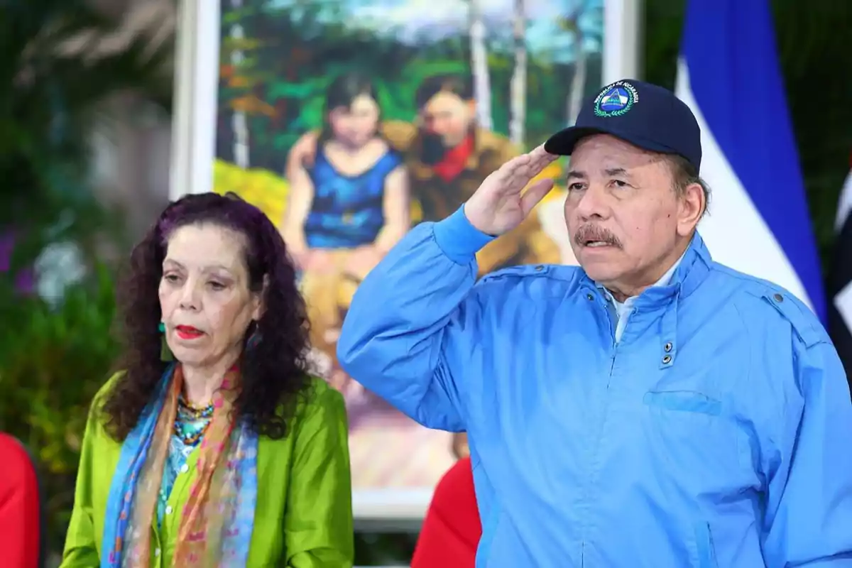 Dos personas de pie, una mujer con ropa colorida y un hombre con chaqueta azul y gorra, frente a una pintura y una bandera.