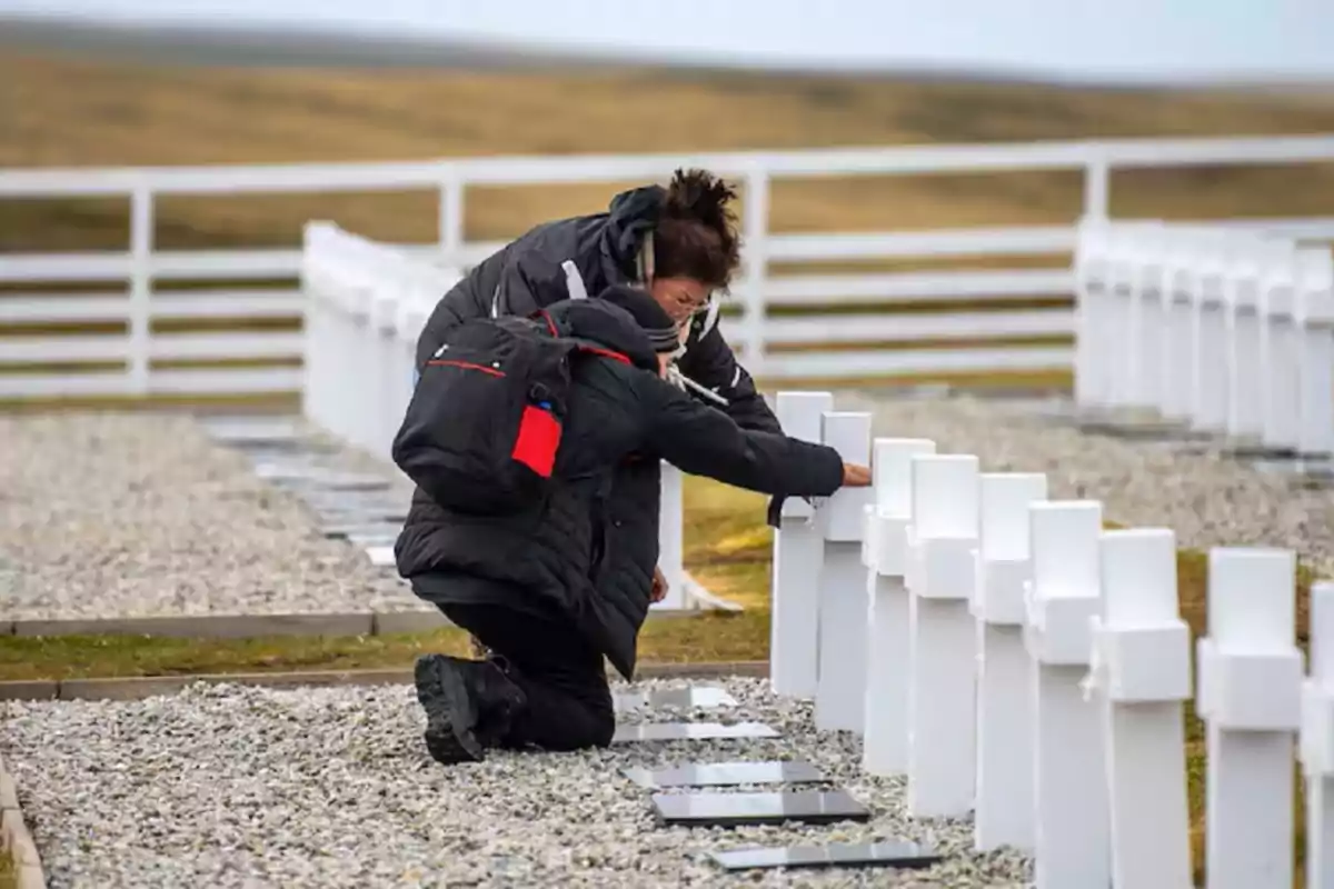 Dos personas vestidas de negro se arrodillan frente a una fila de cruces blancas en un cementerio al aire libre.