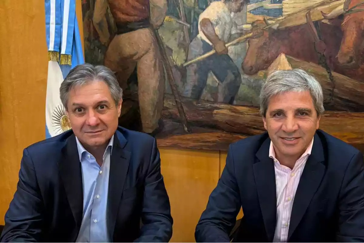 Dos hombres con trajes oscuros sentados frente a una bandera argentina y un mural de fondo.