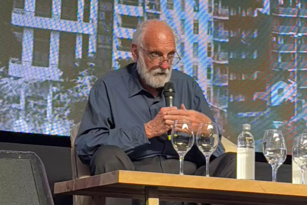 Older man with a beard and glasses speaking into a microphone while sitting on a stage with glasses and a bottle of water on the table.