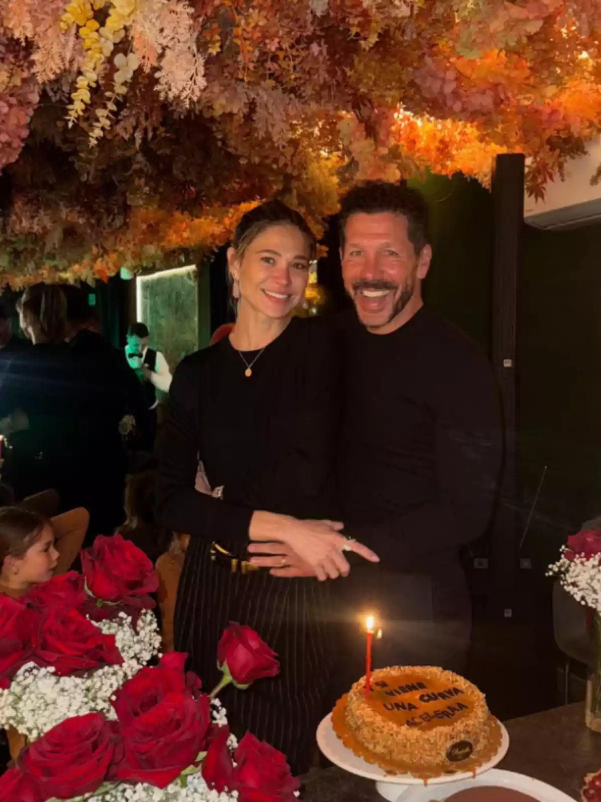 Una pareja sonríe mientras sostiene un cuchillo para cortar un pastel con una vela encendida, rodeados de rosas rojas y un techo decorado con flores secas. Una pareja sonríe mientras sostiene un cuchillo para cortar un pastel con una vela encendida, rodeados de rosas rojas y un techo decorado con flores secas.
