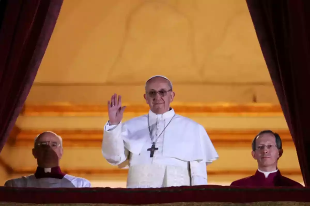 A religious leader dressed in white greets from a balcony accompanied by two people.