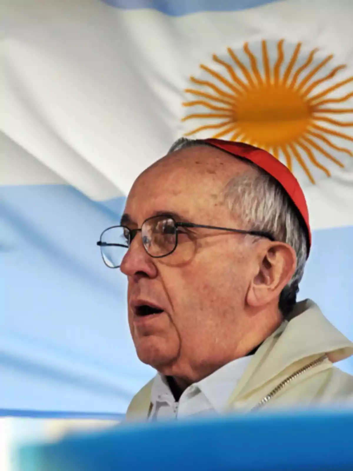 A man with glasses and a red zucchetto in front of an Argentine flag. A man with glasses and a red zucchetto in front of an Argentine flag.