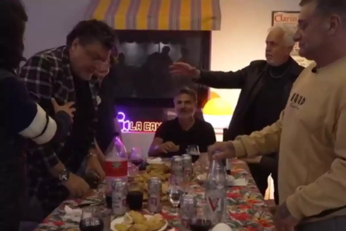 A group of people is gathered around a table covered with a colorful tablecloth, on which there are bottles, cans, and plates of food, as they chat in an informal setting. A group of people is gathered around a table covered with a colorful tablecloth, on which there are bottles, cans, and plates of food, as they chat in an informal setting.