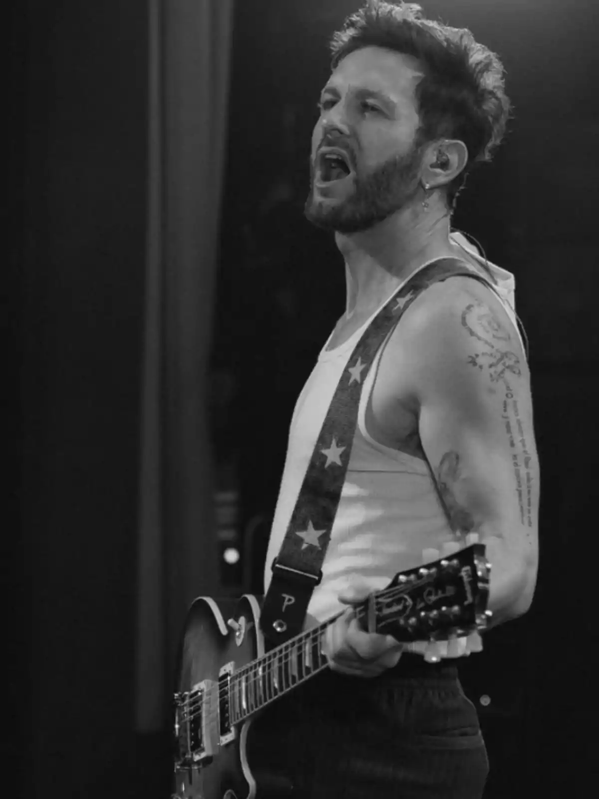 A bearded man playing an electric guitar and singing on stage, wearing a sleeveless jersey and a guitar strap with stars. A bearded man playing an electric guitar and singing on stage, wearing a sleeveless jersey and a guitar strap with stars.
