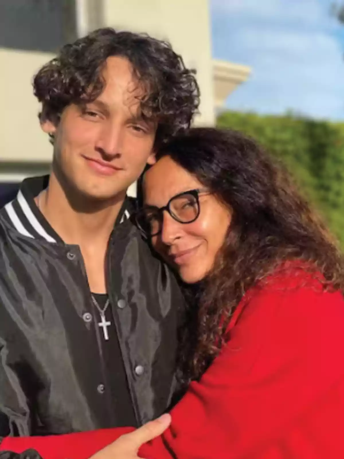 Una mujer con gafas y cabello rizado abraza a un joven de cabello rizado que lleva una chaqueta negra con una cruz colgando del cuello, ambos están al aire libre con un fondo de cielo azul y vegetación. Una mujer con gafas y cabello rizado abraza a un joven de cabello rizado que lleva una chaqueta negra con una cruz colgando del cuello, ambos están al aire libre con un fondo de cielo azul y vegetación.