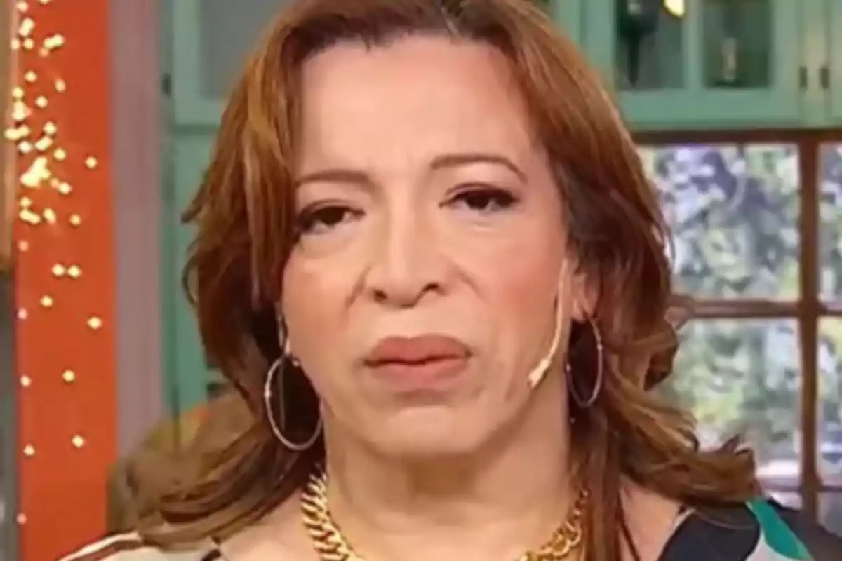 Una persona con cabello castaño y expresión seria, usando aretes grandes y un collar dorado, en un entorno interior con fondo desenfocado. Una persona con cabello castaño y expresión seria, usando aretes grandes y un collar dorado, en un entorno interior con fondo desenfocado.