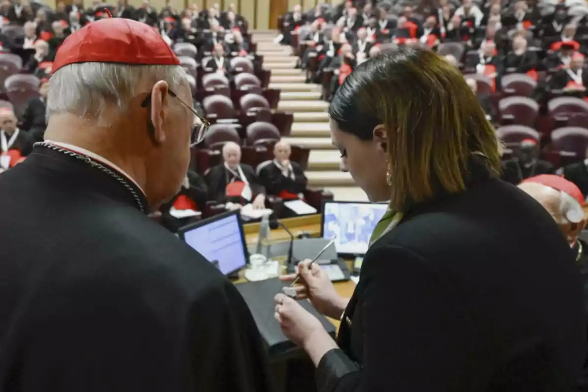 A woman and a clergyman are conversing in a room full of people dressed in formal suits and red caps, while reviewing documents on a table with computers. A woman and a clergyman are conversing in a room full of people dressed in formal suits and red caps, while reviewing documents on a table with computers.