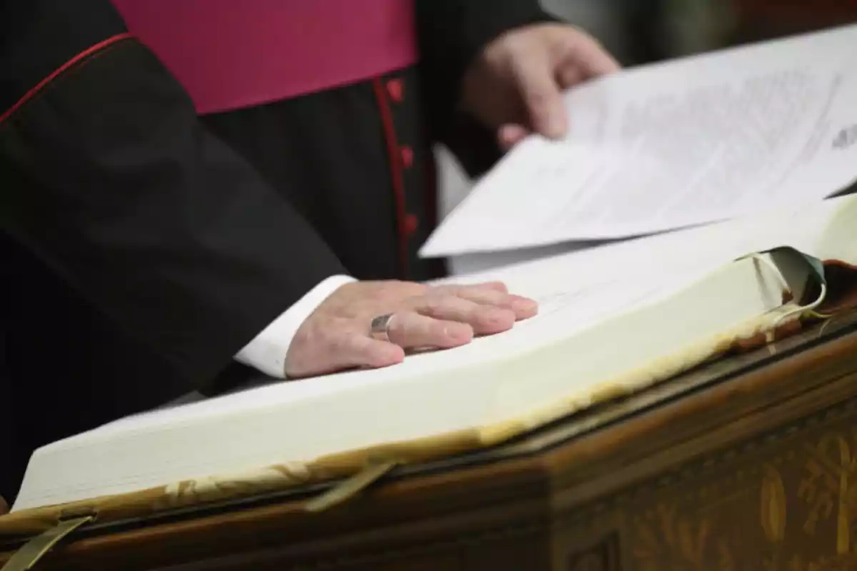 Una persona con vestimenta clerical coloca su mano sobre un libro grande mientras sostiene un documento. Una persona con vestimenta clerical coloca su mano sobre un libro grande mientras sostiene un documento.
