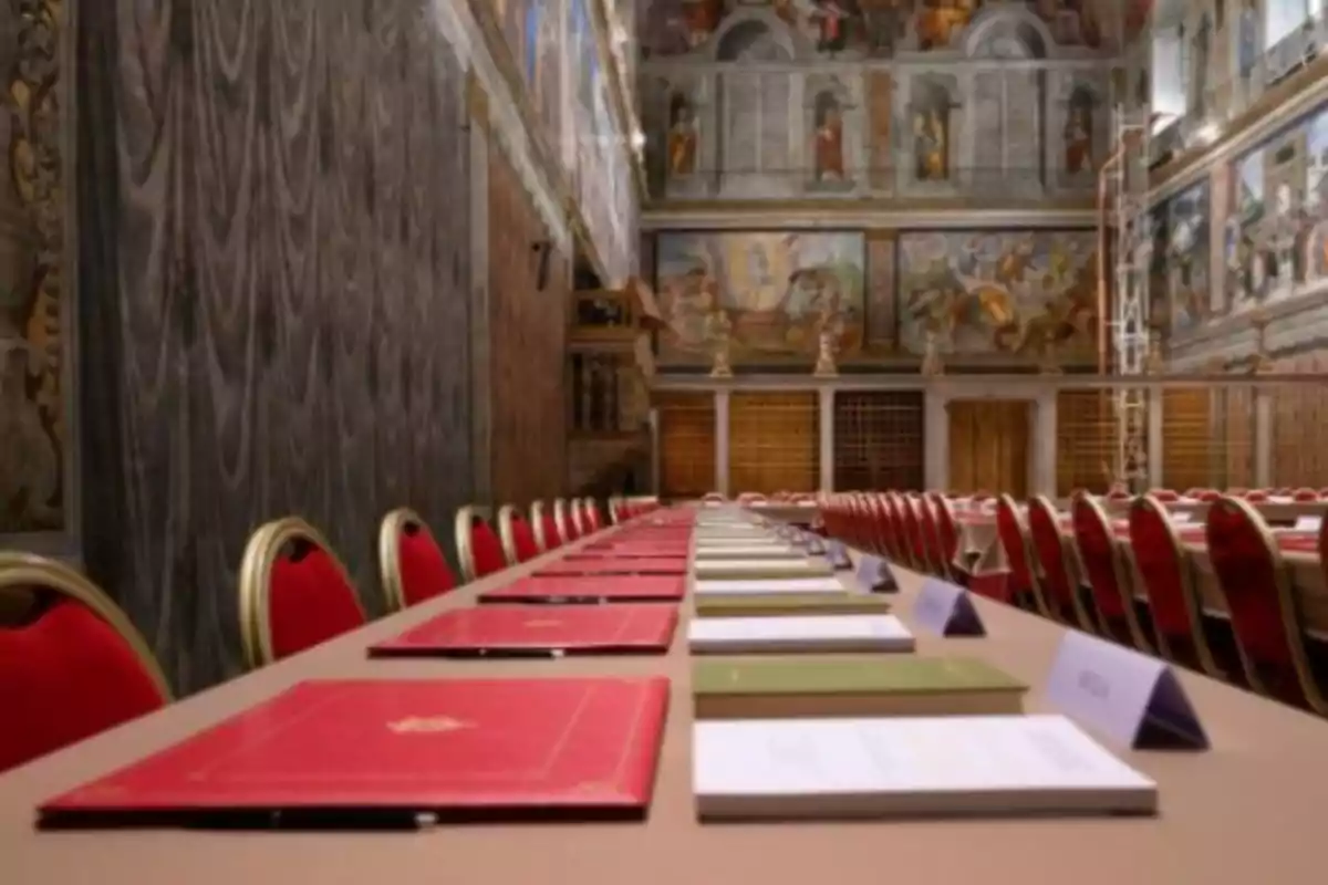 Una larga mesa con sillas rojas y documentos en una sala decorada con frescos en las paredes. Una larga mesa con sillas rojas y documentos en una sala decorada con frescos en las paredes.