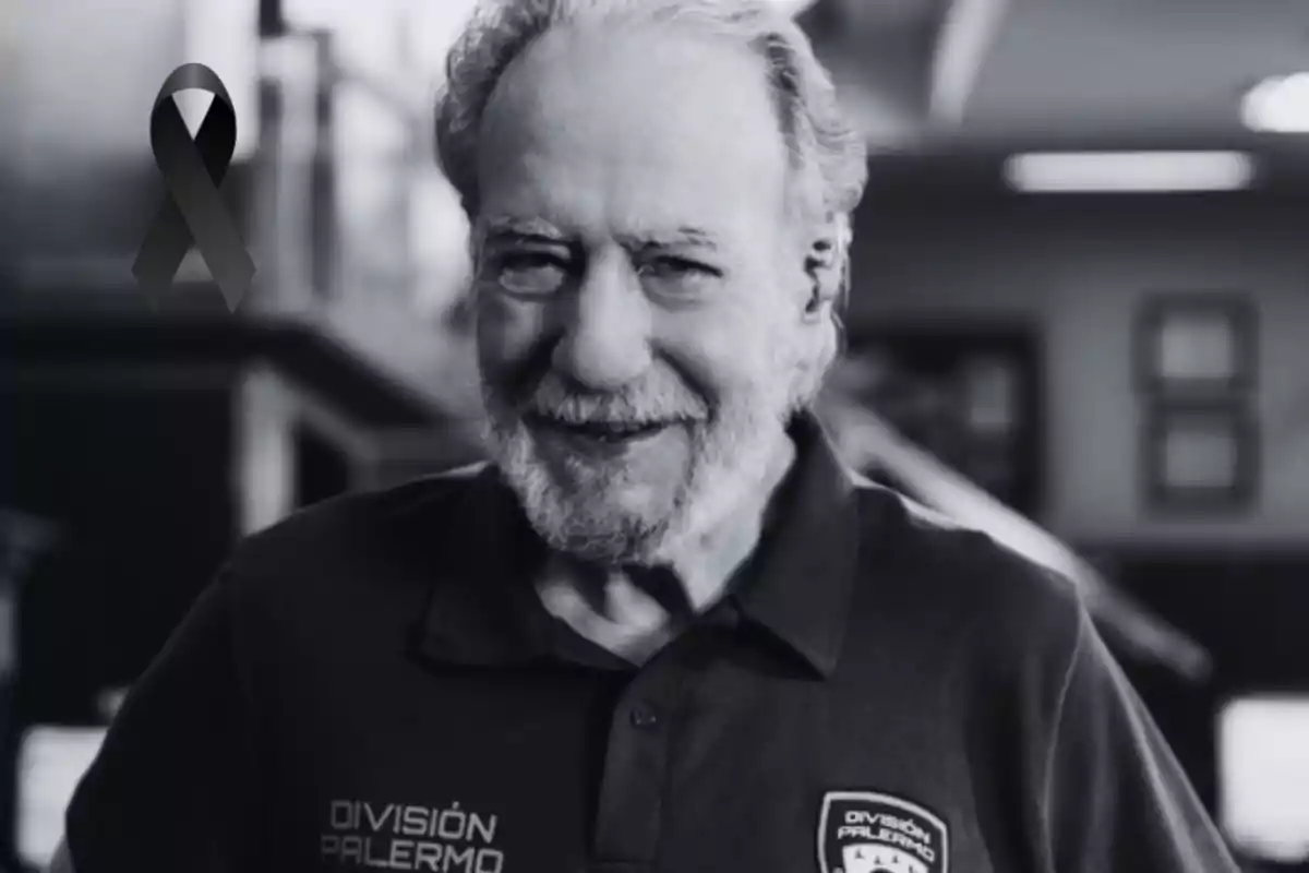 An older man with a gray beard and hair smiles while wearing a t-jersey with the "División Palermo" logo in an indoor setting; in the upper left corner, there is a black ribbon.