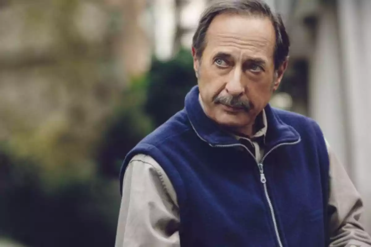 Un hombre con chaleco azul y camisa beige mira hacia un lado en un entorno al aire libre. Un hombre con chaleco azul y camisa beige mira hacia un lado en un entorno al aire libre.