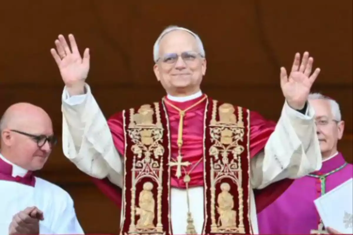 Un hombre vestido con vestimenta religiosa levantando las manos en señal de saludo, acompañado por dos personas.