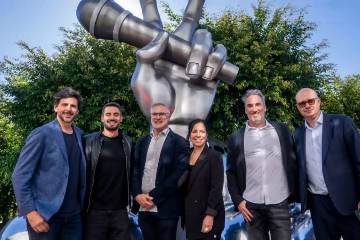 Un grupo de seis personas posando frente a una gran escultura plateada de una mano sosteniendo un micrófono, con árboles verdes de fondo. Un grupo de seis personas posando frente a una gran escultura plateada de una mano sosteniendo un micrófono, con árboles verdes de fondo.