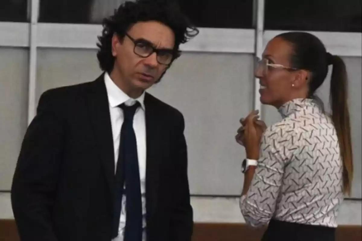 Dos personas conversando en un entorno formal, una de ellas lleva traje y corbata y la otra una blusa estampada. Dos personas conversando en un entorno formal, una de ellas lleva traje y corbata y la otra una blusa estampada.