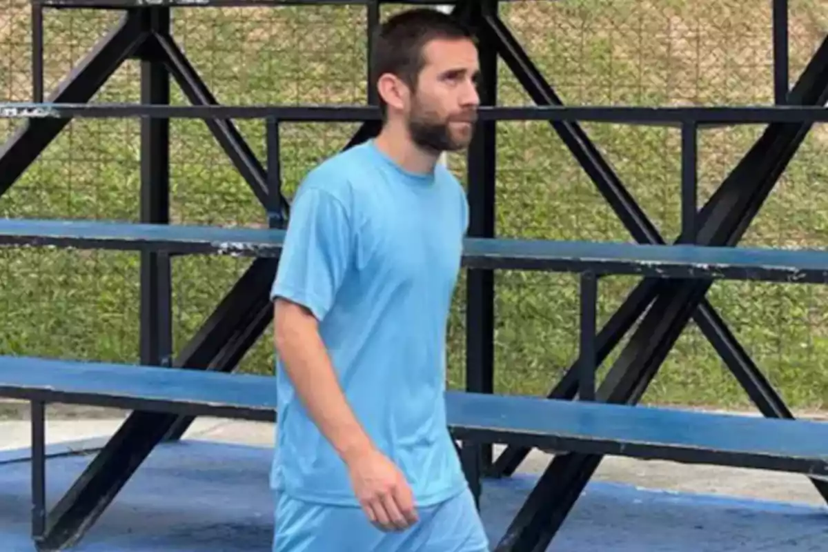 Un hombre con camiseta azul camina frente a unas gradas metálicas en un área al aire libre.