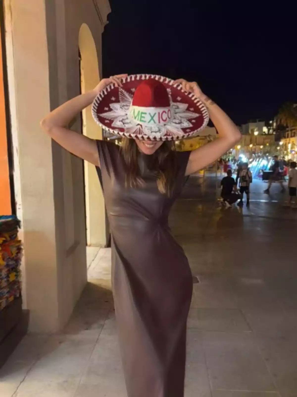 Una persona con un vestido oscuro sostiene un sombrero mexicano que cubre su rostro mientras está de pie en una calle iluminada por la noche. Una persona con un vestido oscuro sostiene un sombrero mexicano que cubre su rostro mientras está de pie en una calle iluminada por la noche.