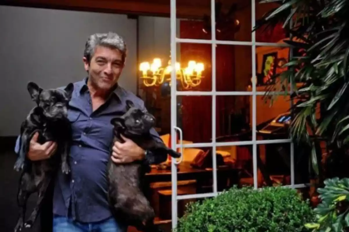 Un hombre sonriente sostiene dos perros pequeños en sus brazos mientras está de pie en la entrada de una casa con una puerta de vidrio abierta y un candelabro encendido en el interior. Un hombre sonriente sostiene dos perros pequeños en sus brazos mientras está de pie en la entrada de una casa con una puerta de vidrio abierta y un candelabro encendido en el interior.