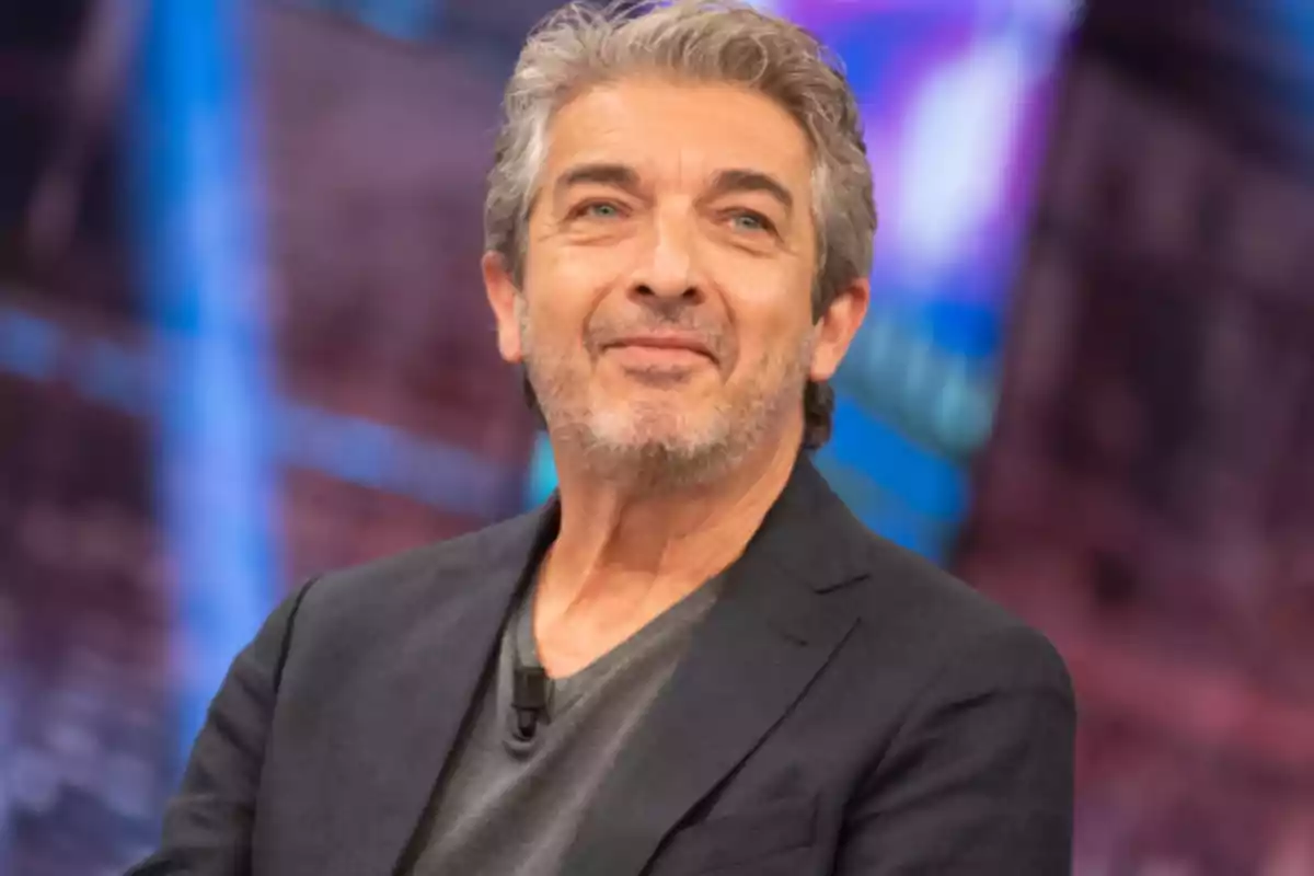 Un hombre de cabello canoso y barba corta viste un saco oscuro y una camiseta gris mientras sonríe ligeramente en un entorno con luces de colores desenfocadas.