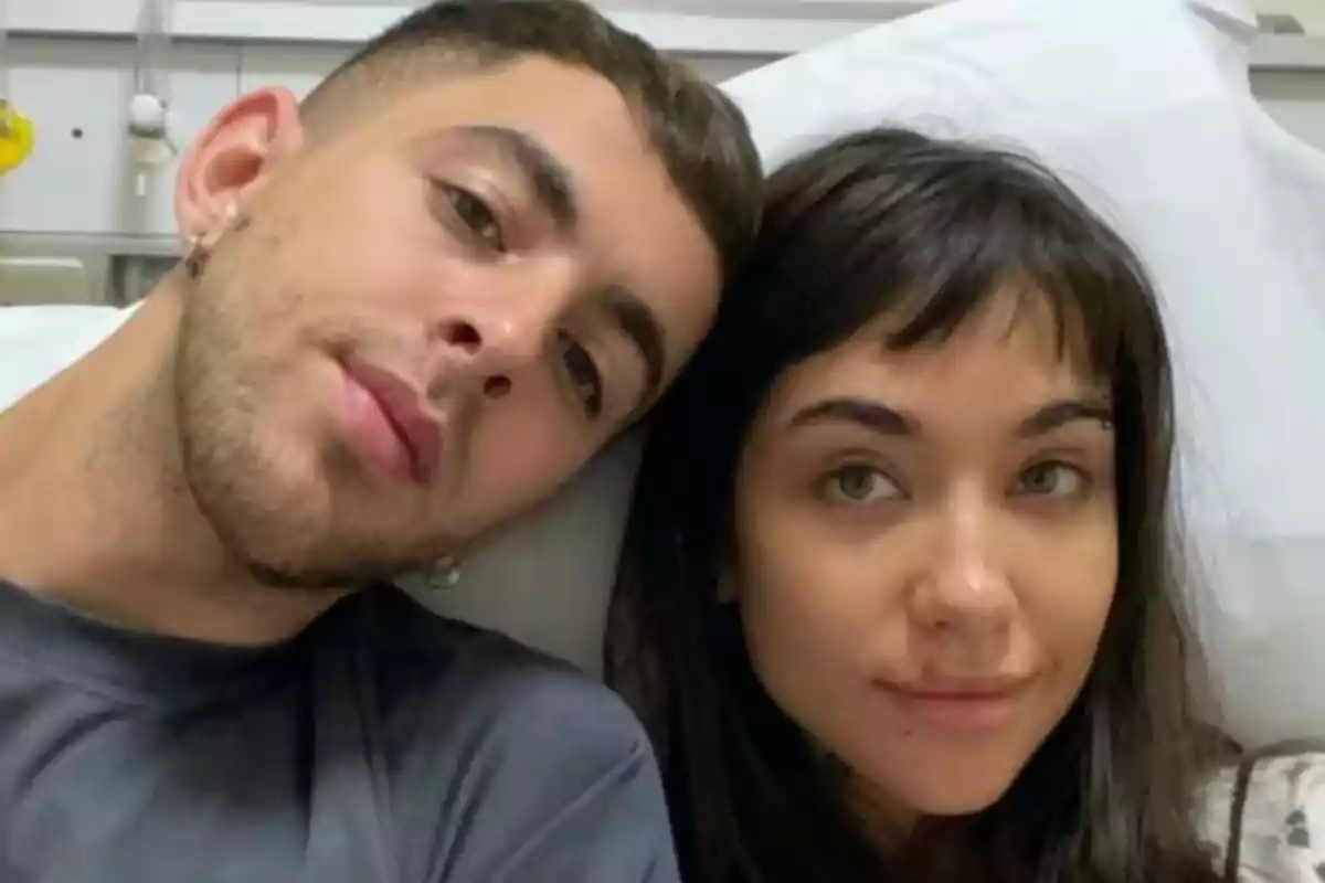 Dos personas recostadas juntas, mirando a la cámara, en un entorno que parece ser un hospital.