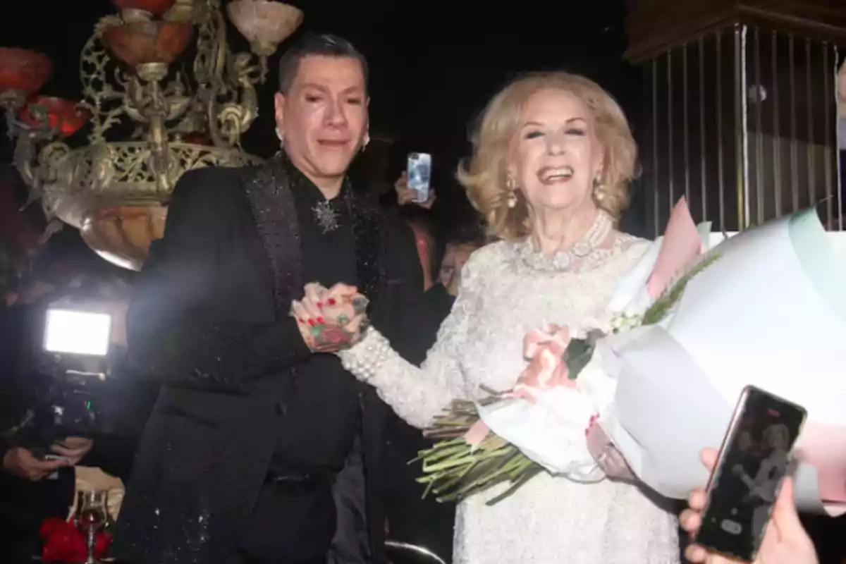 Two smiling people pose together at a fancy event, one of them holding a bouquet of flowers, and there is a decorative chandelier in the background. Two smiling people pose together at a fancy event, one of them holding a bouquet of flowers, and there is a decorative chandelier in the background.