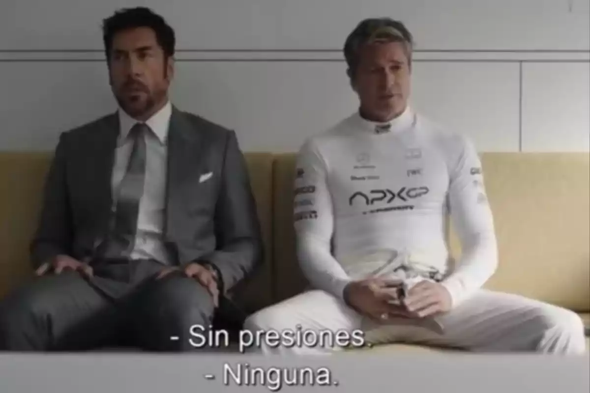 Two men sitting on a couch, one in a gray suit and the other in a white racing suit, with subtitles that say Two men sitting on a couch, one in a gray suit and the other in a white racing suit, with subtitles that say