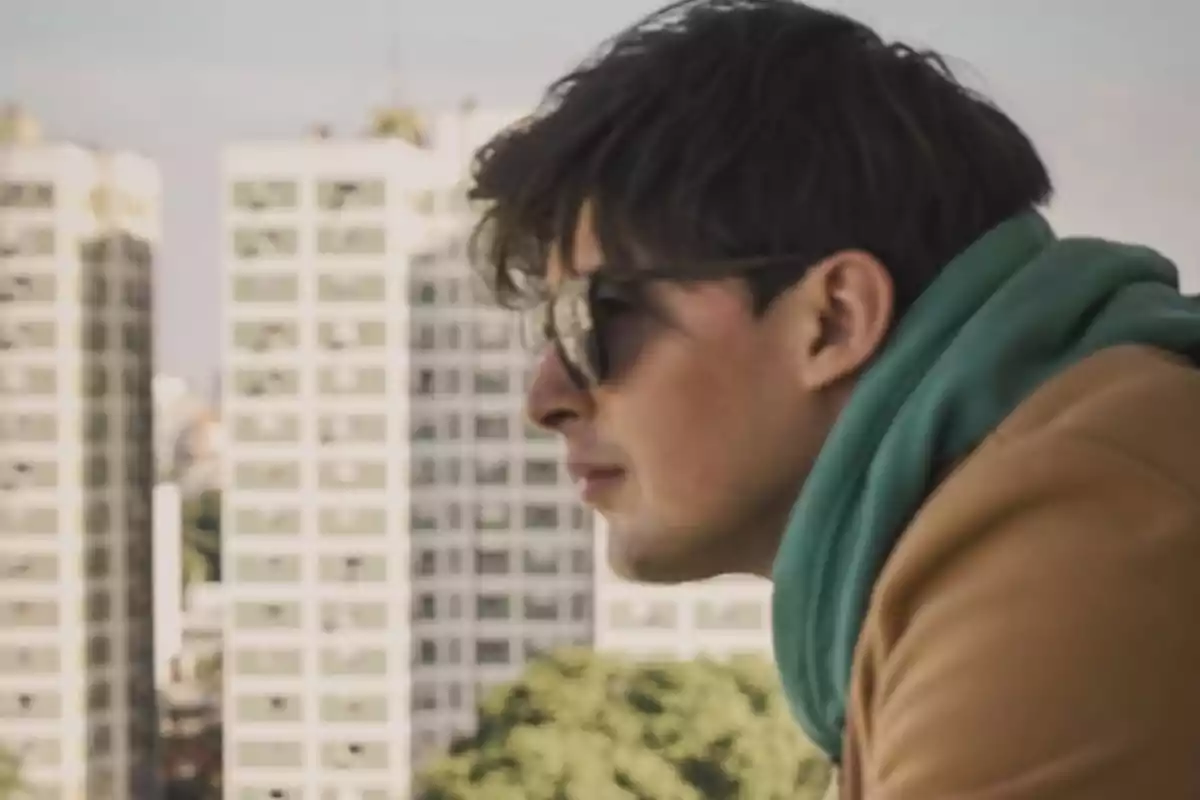 Un hombre con gafas de sol y chaqueta marrón mira pensativo hacia el horizonte con edificios de fondo. Un hombre con gafas de sol y chaqueta marrón mira pensativo hacia el horizonte con edificios de fondo.