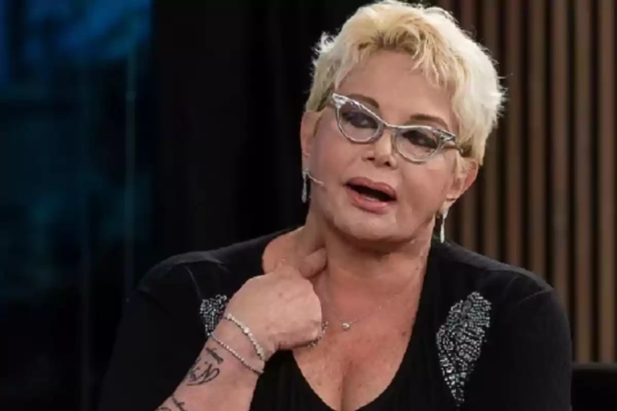 A person with short blonde hair and glasses, dressed in black, is talking while touching their neck.